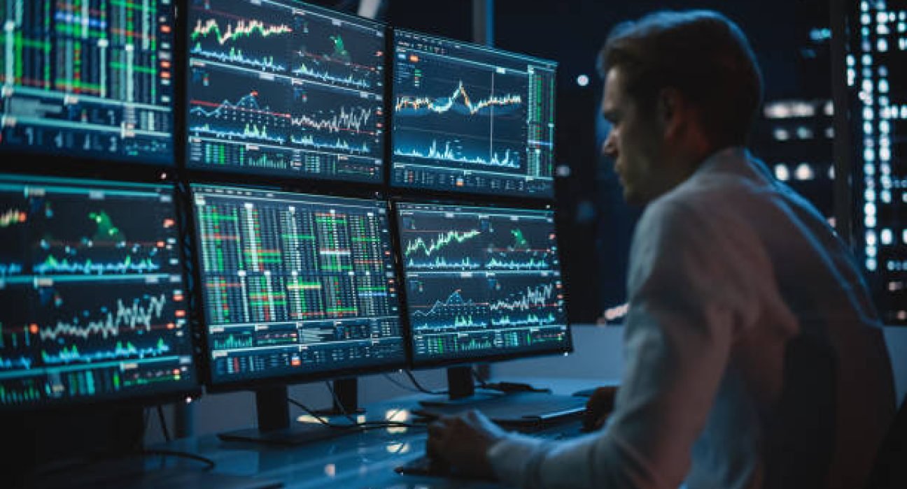Financial Analyst Working on a Computer with Multi-Monitor Workstation with Real-Time Stocks, Commodities and Exchange Market Charts. Businessman Works in Investment Bank Downtown Office at Night.